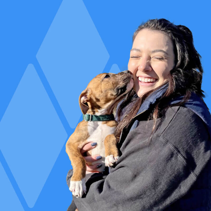 image description: girl standing in black jacket smiling while puppy licks her cheek